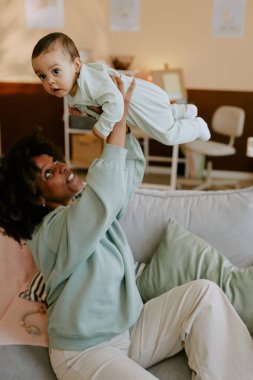 Genç Afrikalı Amerikalı anne, bebeğini hafif bir bebek odasında yumuşak koltukta otururken kaldırıyor.