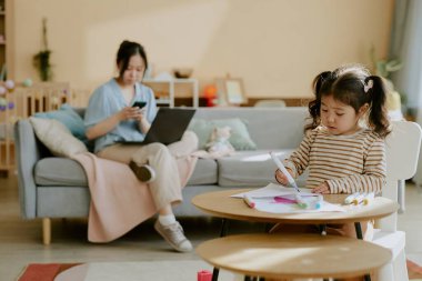 Asyalı küçük kız çizim becerisini geliştirirken annesi de arka plandaki bulanık telefonu kontrol ediyor.