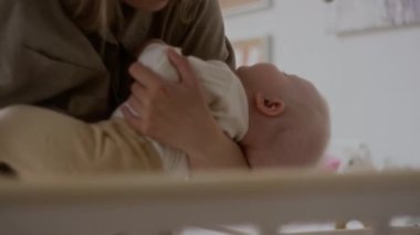 Medium shot of unrecognizable young mother softly and securely placing baby into crib leaving little child safely sleeping on white pillow