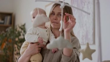 Orta ölçekli gülümseyen beyaz kadın yeni şekil ve dokular sunarak bebek kızının yumuşak bulutlar ve yıldızlarla hareket etmesine dikkat çekiyor.