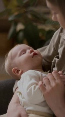 Annesinin kollarında uyuyan sevimli beyaz bebeğin dikey yakın çekimi. Masum küçük çocuğuna hayranlık duyuyor. Sevecen bir şekilde sevimli yüzüne bakıyor ve minik elini tutuyor.
