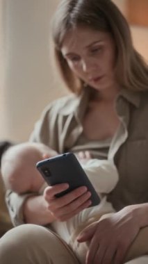 Beyaz bir kadının akıllı telefon kullanırken bebeğini emzirirken kollarında huzur içinde uyuduğu dikey bir fotoğraf.