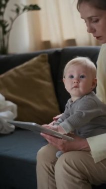 Kanepede oturan beyaz bir kadının tablet ekran çizimine odaklanmış meraklı bir bebeği tutarken ya da güzel motor ve duyusal beceriler geliştirerek oyun oynarken dikey orta pozu.