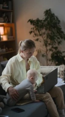 Dikey çekimde, genç beyaz kadın kanepeye oturmuş laptopuyla bebeğinin bakımını merakla oynarken ve yakınlarda oynaşırken iş birliği yapmaya çalışıyor.