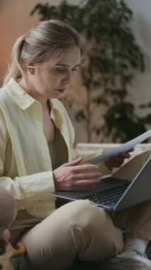 Küçük oğlu ya da kızı yakınlardaki tahta oyuncakla oynarken kucağında dizüstü bilgisayarla kanepede oturup bacak bacak bacak üstüne atmış genç beyaz bir annenin dikey pozu.