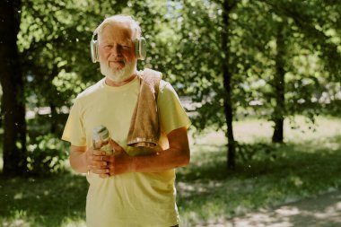 Gümüş kablosuz kulaklık takan ve fotoğraf için poz verirken elinde bir şişe su tutan kendine güvenen bir adam.