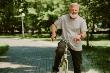 Bisiklet sürdükten sonra mola veren gümüş saçlı adam doğrudan kameraya bakıyor ve fotoğraf için poz veriyor.