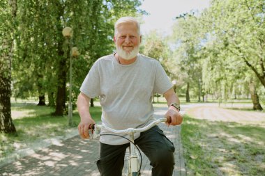 Günlük giysiler içinde gülümseyen gümüş saçlı adamın portresi, parkta bisiklet sürüyor.