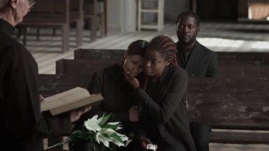 Siyahi kadın, kilisedeki ahşap bankta oturmuş elinde İncil 'le dua eden Afrikalı Amerikalı bir adamın arka planda oturmasını dinlerken beyaz kadın arkadaşını teselli ediyor.