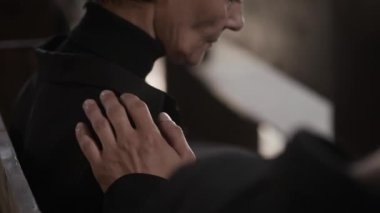 Kilisedeki cenaze töreninde yanında oturan kederli kadının elini omzuna koyan tanınmayan bir adam onun eline dokunuyor.