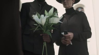 Beyaz çiçekli ve mendilli üç aile ferdi açık hava cenaze töreninde yan yana dururlar.