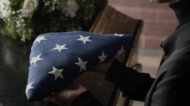 Kilisedeki anma töreninde beyaz çiçeklerin yanındaki tabut kapağına üçgen şeklinde Amerikan bayrağını koyan tanınmayan bir kadının yakın plan fotoğrafı.