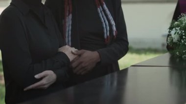 Tanımlanamayan iki bayan arkadaş, tabutun başında ellerinde İncil ve açık hava cenaze töreninde dua okuyan rahibin yanında duruyor.