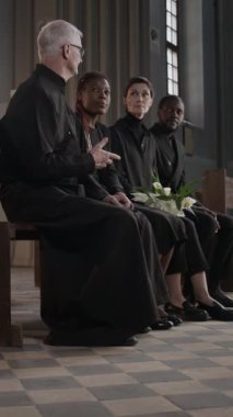 Baş papazın, siyah takım elbiseli üç aile üyesiyle kilisedeki ahşap sıralarda otururken görüntüsü.