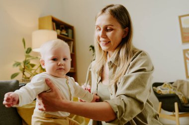 Anne bebeğiyle meşgul, rahat, aydınlık bir oturma odasında otururken kucağında gülümsüyor ve doğal dekorları ve mobilyaları var.