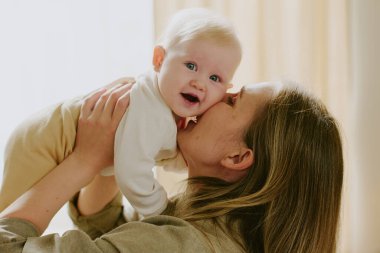 Neşeli anne mutlu bebeğini yanağından kaldırıp öpüyor, şefkatli ve sevgi dolu bir anı yakalıyor. Doğal ışık, anne ve çocuk arasındaki duygusal bağı güçlendirir.