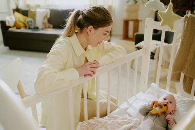 Gülümseyen anne, bebeğin etrafında yumuşak ışıklandırma ve pelüş oyuncaklarla bebeğine bakıyor. Sahne ayrıca arka planda rahat bir yaşam alanı içerir