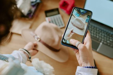 Akıllı telefon tutan kişi, bej şapka görüntüsü alıyor, hesap makinesi ve dizüstü bilgisayarla birlikte, ayrıntılı tokaya odaklanıyor. Sahne sıcak, yakın düzenlenmiş çalışma alanında ayarlandı