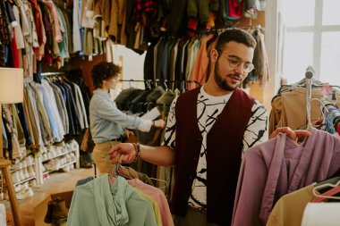Erkek modern butikte giysileri dikkatle seçerken konsantrasyonlu giysi raflarına katlanmış giysiler ve arka planda gezinen kadınlar koyuyor.