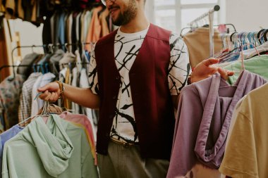 Gülümseyen adam canlı ikinci el mağazasında kıyafet karıştırıyor, çeşitli renklerde elbise askıları tutuyor, alışveriş deneyimleriyle meşgul, çeşitli moda eşyalarıyla çevrili.