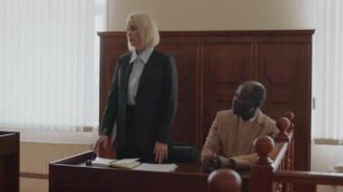 Beyaz kadın avukat, mahkeme sırasında Afrika kökenli Amerikalı erkek davacının yanındaki ahşap masada otururken iddianame konuşması yapıyor.