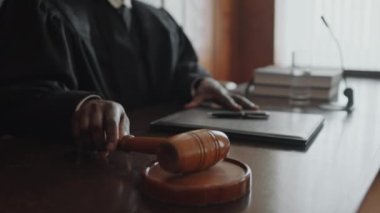Siyahi erkek yargıcın tahta tokmakla ses bloğuna vuruşunu, klasörün açılışını ve mahkemenin başlamasını görüntüle.