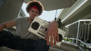 Genç rapçinin elinde kırmızı bandana ve sokak kıyafetleriyle kentin üst geçidinin altında merdivenlerde otururken hip hop şarkıları söylerken, gün ışığının kameraya yansıdığı görüntüyü büyüt.