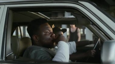 Ciddi Afrikalı Amerikalı bir adam arabada arkadaşının yanında oturuyor, sigara içiyor ve erkek arkadaşını selamlıyor.