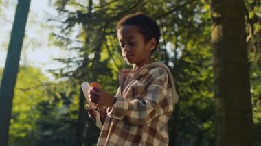 Güneşli yaz gününde yemyeşil ormanlarda büyüteç kullanarak taze mantarları iyice inceleyen meraklı Afrikalı genç çocuğun orta boy fotoğrafı.