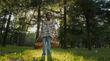 Ormanda elinde hasır sepetlerle dikilen ve güneşli bir günde mantar toplamaya hazırlanırken kameraya poz veren Afro-Amerikan bir çocuğun portresi.