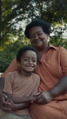 Sıcak bir yaz günü bankta oturmuş kameraya poz verirken gülümseyen küçük çocuğu kucaklayan yaşlı Afrikalı Amerikalı kadının dikey portresi.
