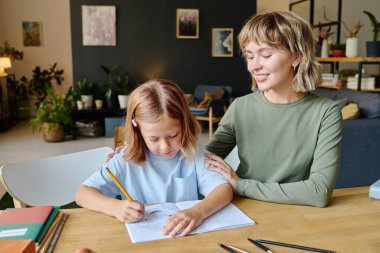 Sarışın anne okul ödevi yazarken kızına destek oluyor.