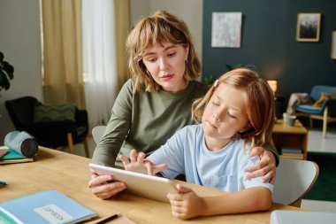 Genç beyaz anne kızıyla masada oturuyor ve tabletle resim çizerken ona sarılıyor.