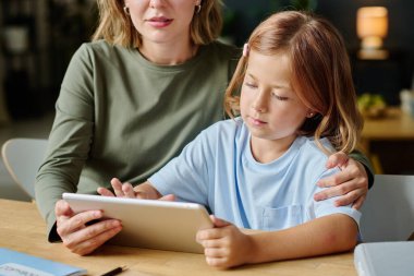 Annesinin ona sarılırken tablet kullanan sevimli kız akıllı saç kesiminin orta boy görüntüsü.