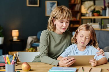 Anne ve kızı benzer saç kesimleriyle ahşap masada oturup tabletle çizgi film izliyorlar.