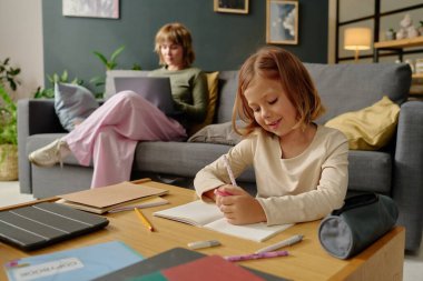 Annesi bulanık arka planda çalışırken küçük kız yerde oturup renkli kalemlerle resim çiziyor.