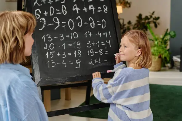 stock image Proud girl holding white chalk and doing sums while looking at her mom