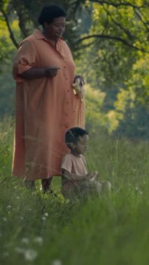 Yeşil parktaki nehir kıyısında oturan Afrikalı Amerikalı çocuğun tam dikey görüntüsü. Büyükannesinin yardımıyla ördekleri besliyor.