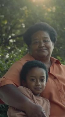 Yazın parkta bankta oturan Afrikalı Amerikalı büyükannenin dikey portresi. Küçük torununu kucaklıyor, gülümsüyor ve kameraya bakıyor.