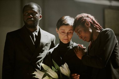 Mournful woman in total black clothes holding madonna lilies surrounded by her African American friends who supporting her and crying clipart