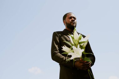 Siyah kostümlü, beyaz zambak buketi tutan ve kaybından yakınan Afro-Amerikan bir adamın düşük açılı çekimi.