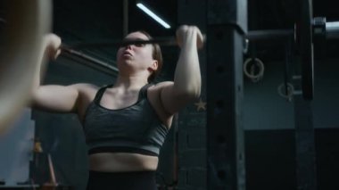 Güçlü genç bayan sporcu, üstten kaldırma hareketi yapıyor, sonra halteri askıya alıp dinleniyor.
