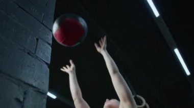 Güçlü genç bir kadın duvar topu çömelmesi yapıyor, sonra da spor yaparken topu yere bırakıp gidiyor.