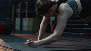 Genç sporcu kadın tahtada duruyor, dirseklerine yaslanıyor ve başlangıç pozisyonu alıyor, sonra mindere uzanıyor ve spor yaparken dinleniyor.