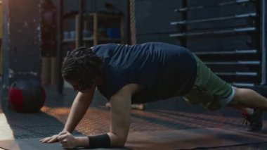 Genç erkek sporcu kalas egzersizi yapıyor. Dirseklerden başlayıp ellere doğru yükseliyor. Sonra da spor salonunda paspasın üzerine oturuyor.