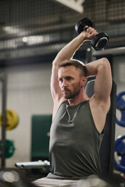 Olgun odaklı adam bankta oturuyor ve ağır kauçuk halterle omuz egzersizi yapıyor.