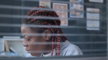 Camdan aşağı doğru eğil. Yoğunlaştırılmış Afro-Amerikan kadın doktorların hasta bilgilerini not defterine yazıp sonra da iş yerindeki bilgisayarını kullanarak kameraya çek.