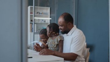 Afrikalı Amerikalı bir baba kucağında küçük kızıyla resim çiziyor ve not tutan kadın bir çocuk doktoruyla konuşuyor.