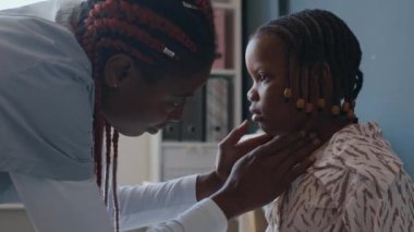 Çocuk kliniğinde boğazını dikkatle kontrol ederken Afrikalı Amerikalı kadın hekimin küçük bir hastanın boynuna dokunuşunu yakınlaştırın.
