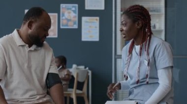 Kadın hekimin, klinikteki aile randevusunda monitörle ölçerken erkek tansiyonu hakkında yorum yaparken, arka planda oynayan iki kız görüntüsünü yukarı kaldır.